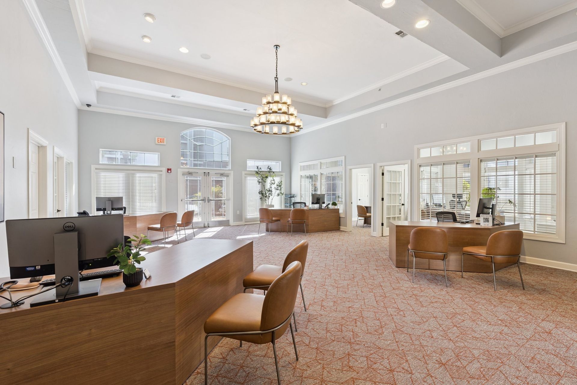 Reception area with light-colored walls, tan desks, chairs, and speckled flooring. Windows offer natural light.