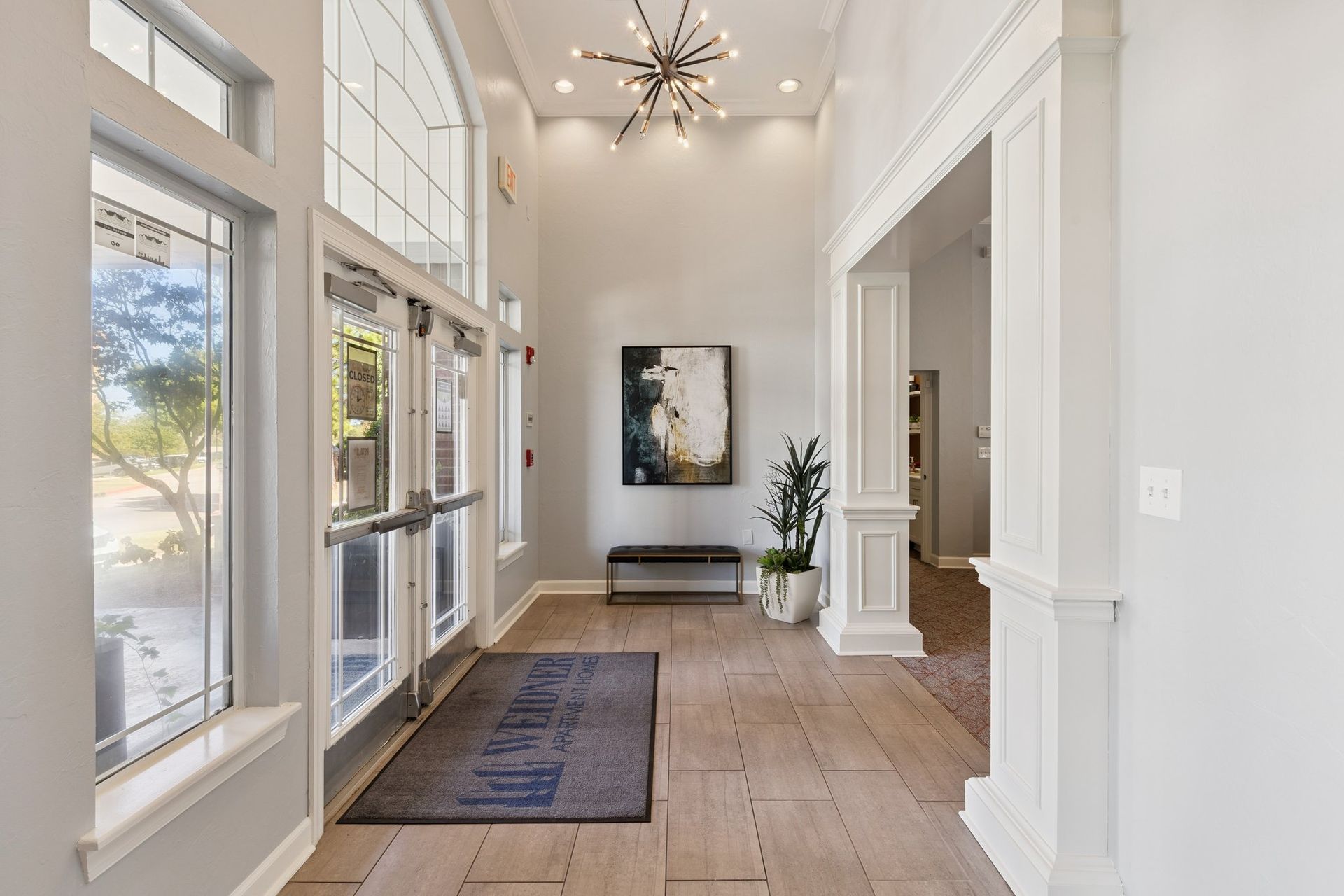 Lobby with glass doors, tall windows, and a modern chandelier. There's a bench, art, and a potted plant.