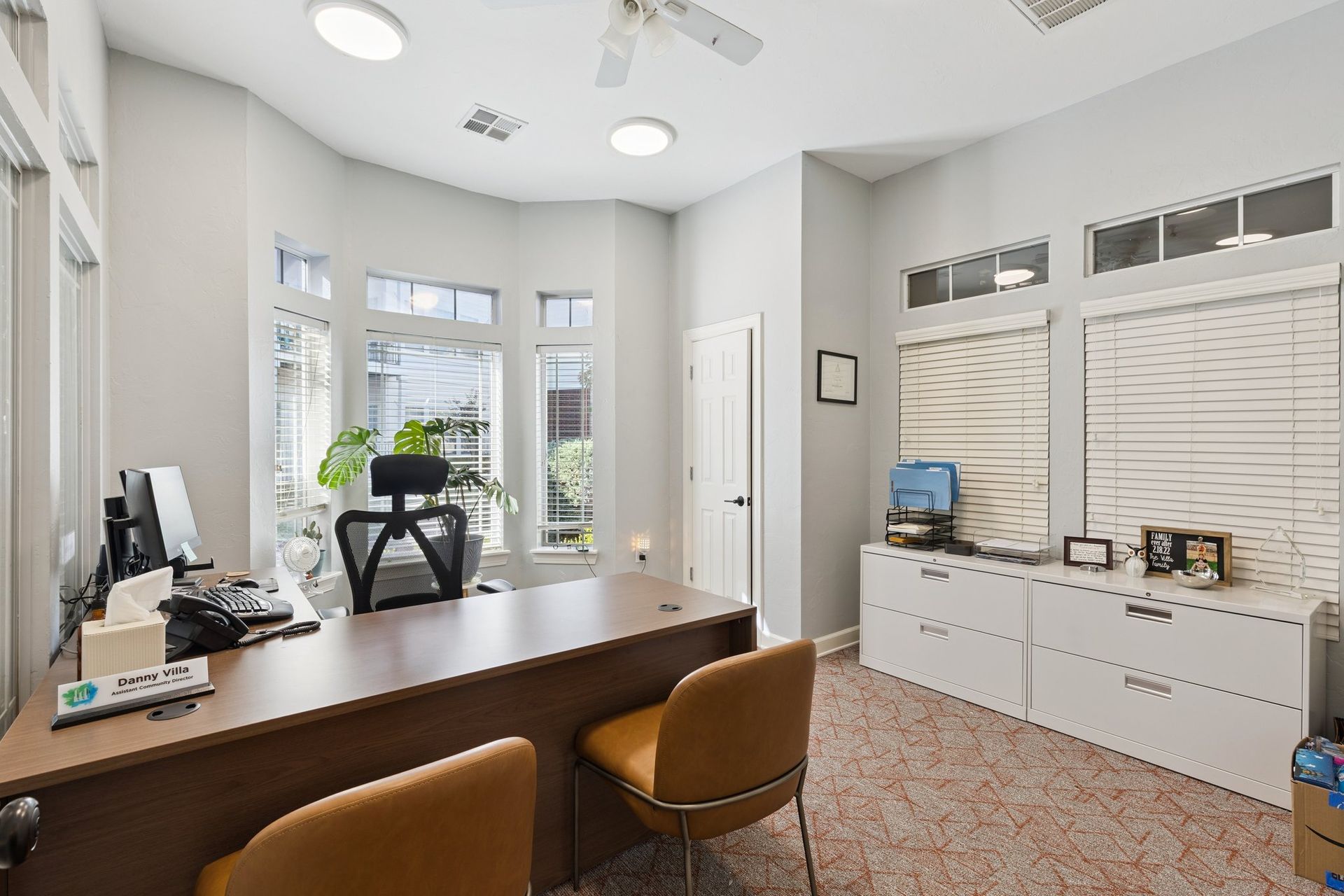 Office with a desk, chair, and large windows. Beige walls, white cabinets, and tan carpet.