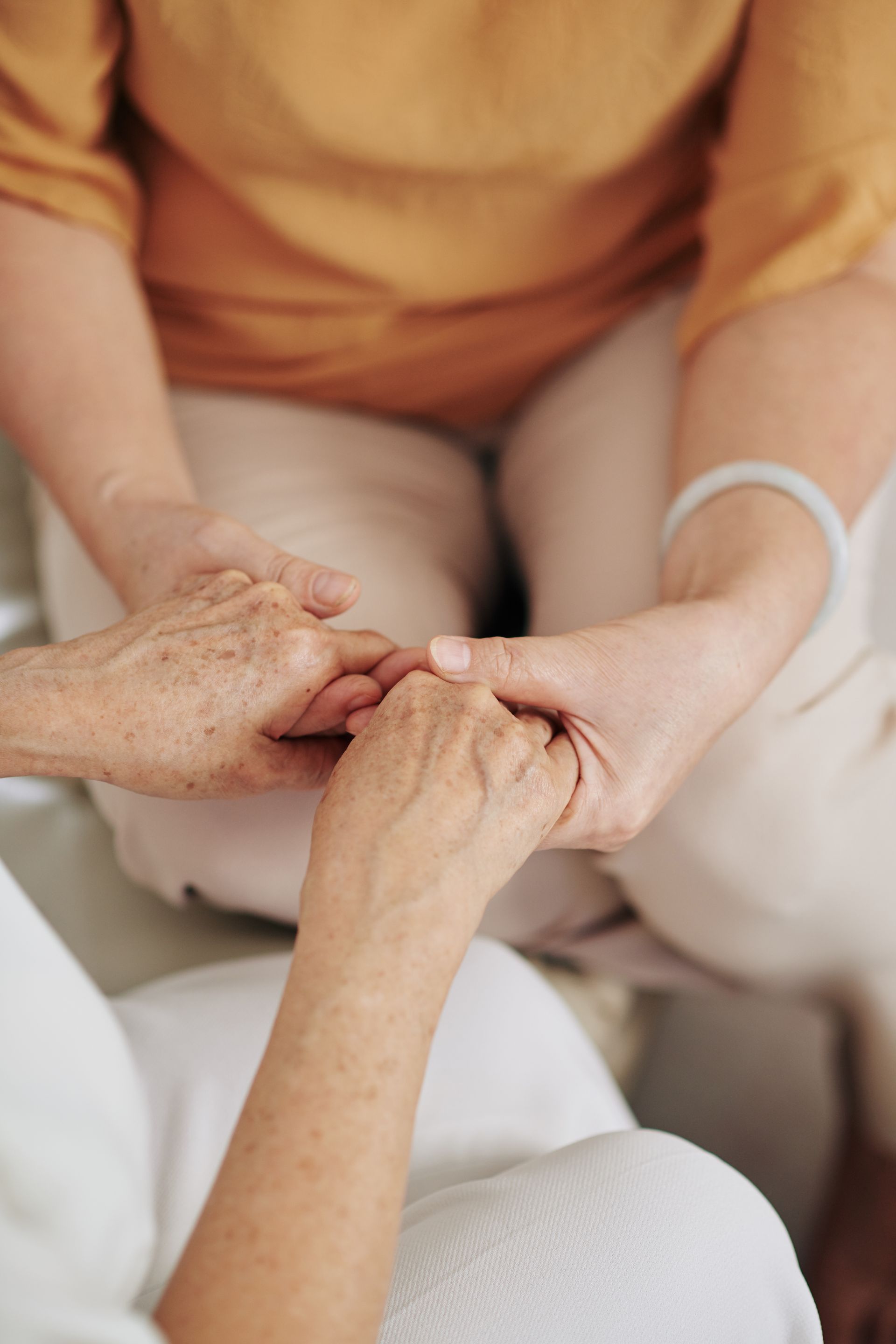 Two people holding hands, one with aged skin, seated, warm tones.