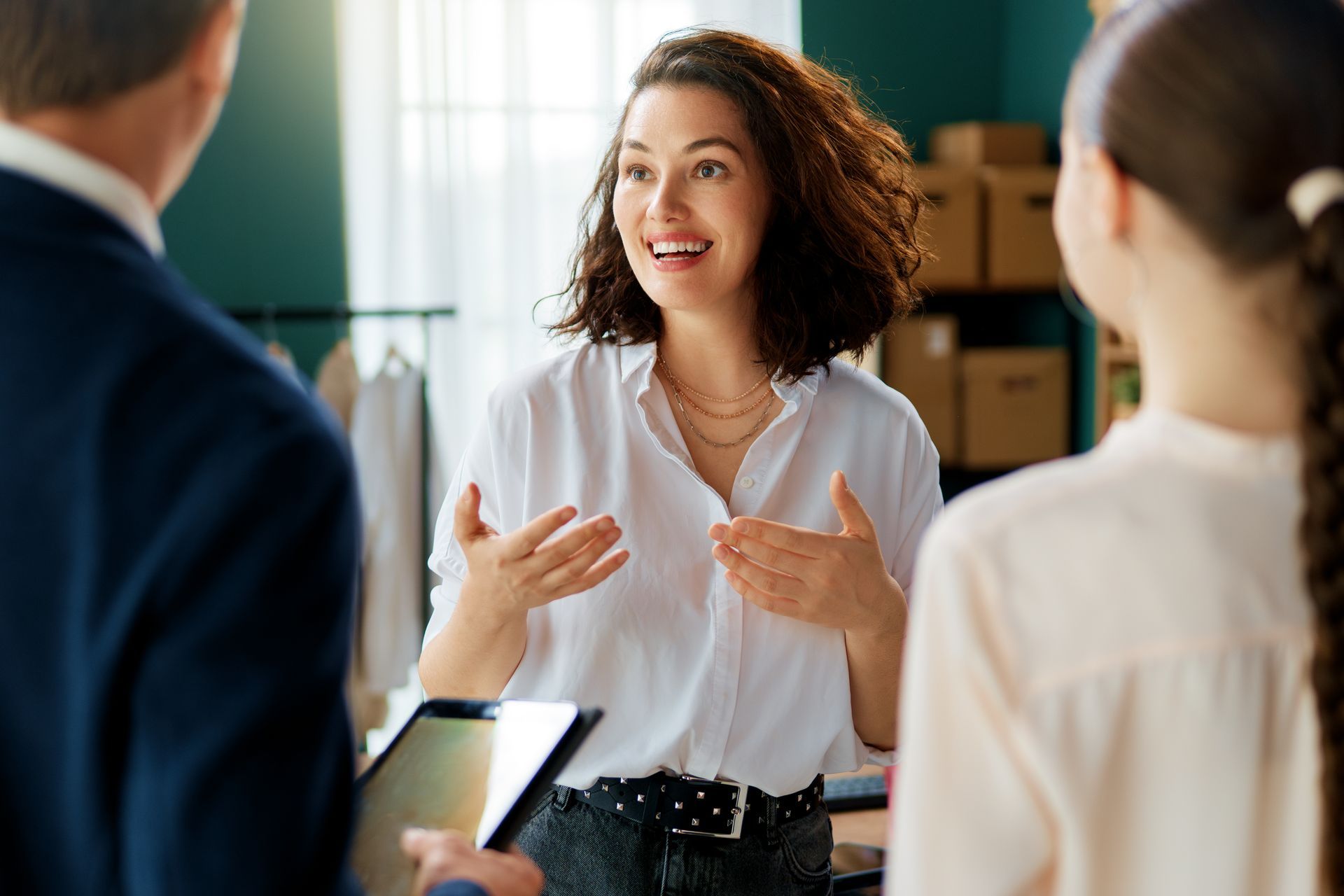 Woman gesturing, talking to two people, in an office space. She has brown hair, a white shirt. The others are out of focus.