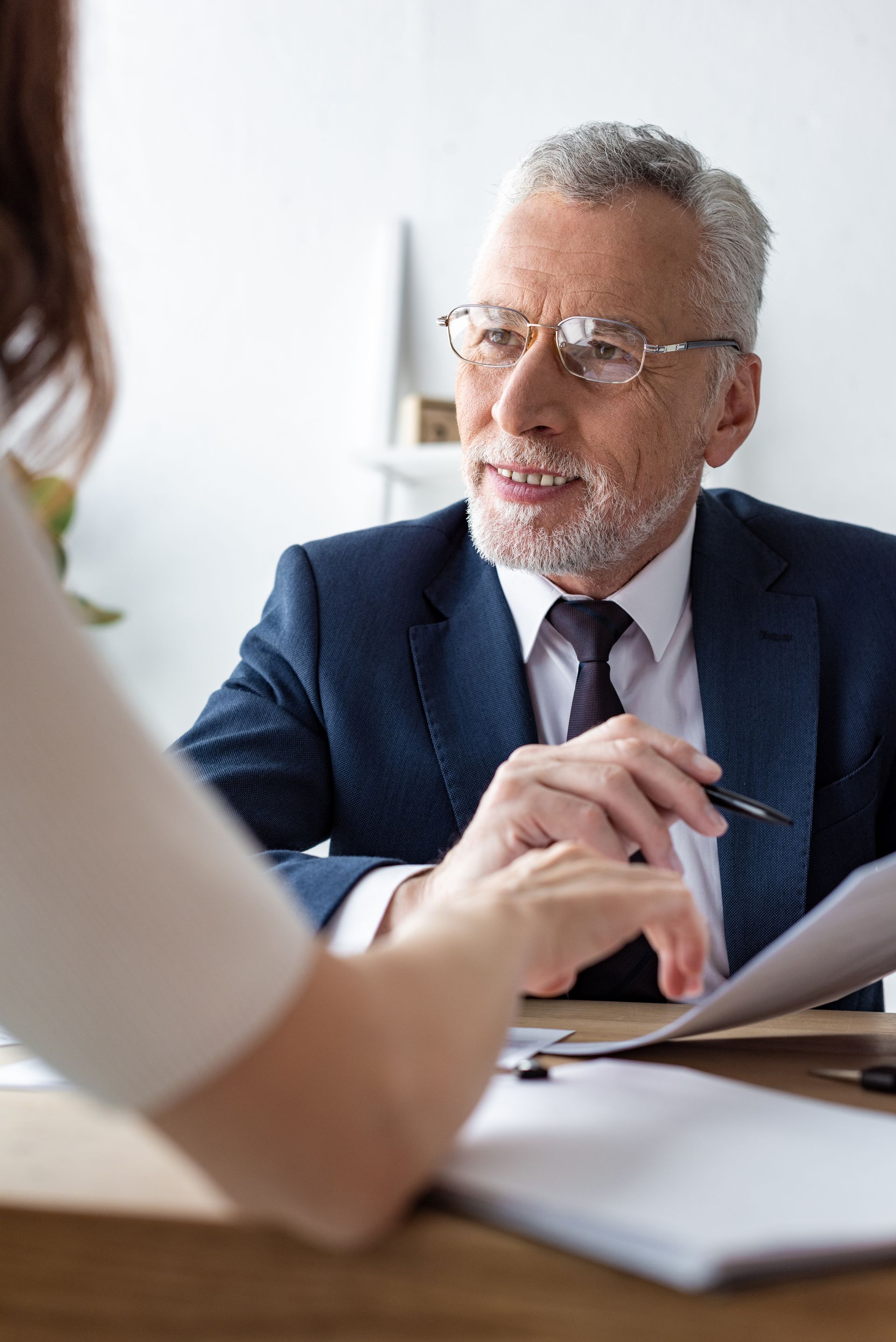 Senior man in suit smiles while reviewing documents with a woman at a desk.