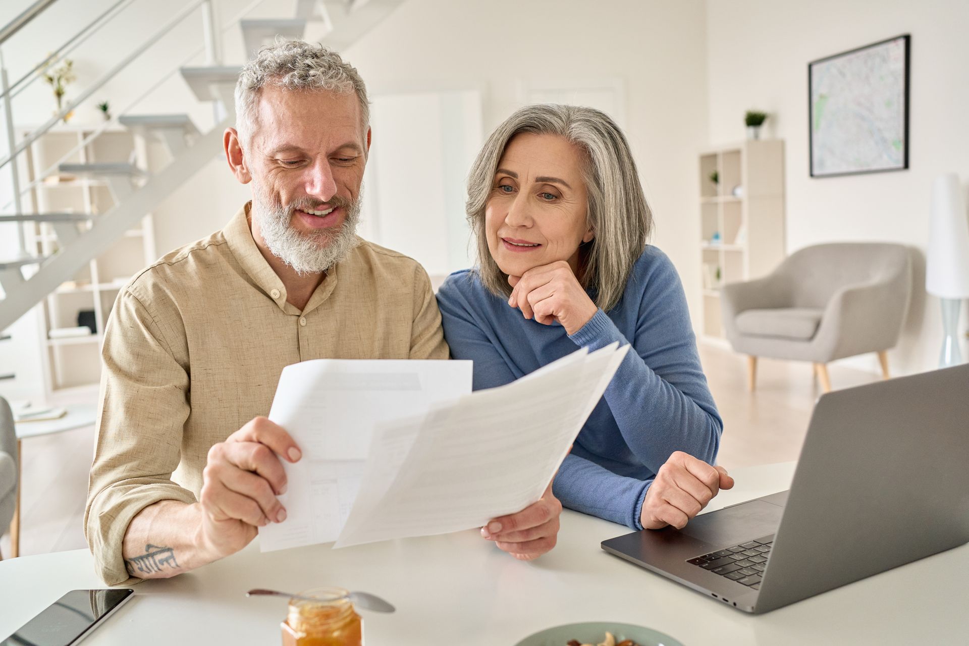 Older couple reviewing paperwork together at a table with a laptop in a bright home.