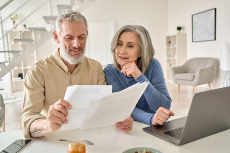 Older couple reviewing paperwork together at a table with a laptop in a bright home.