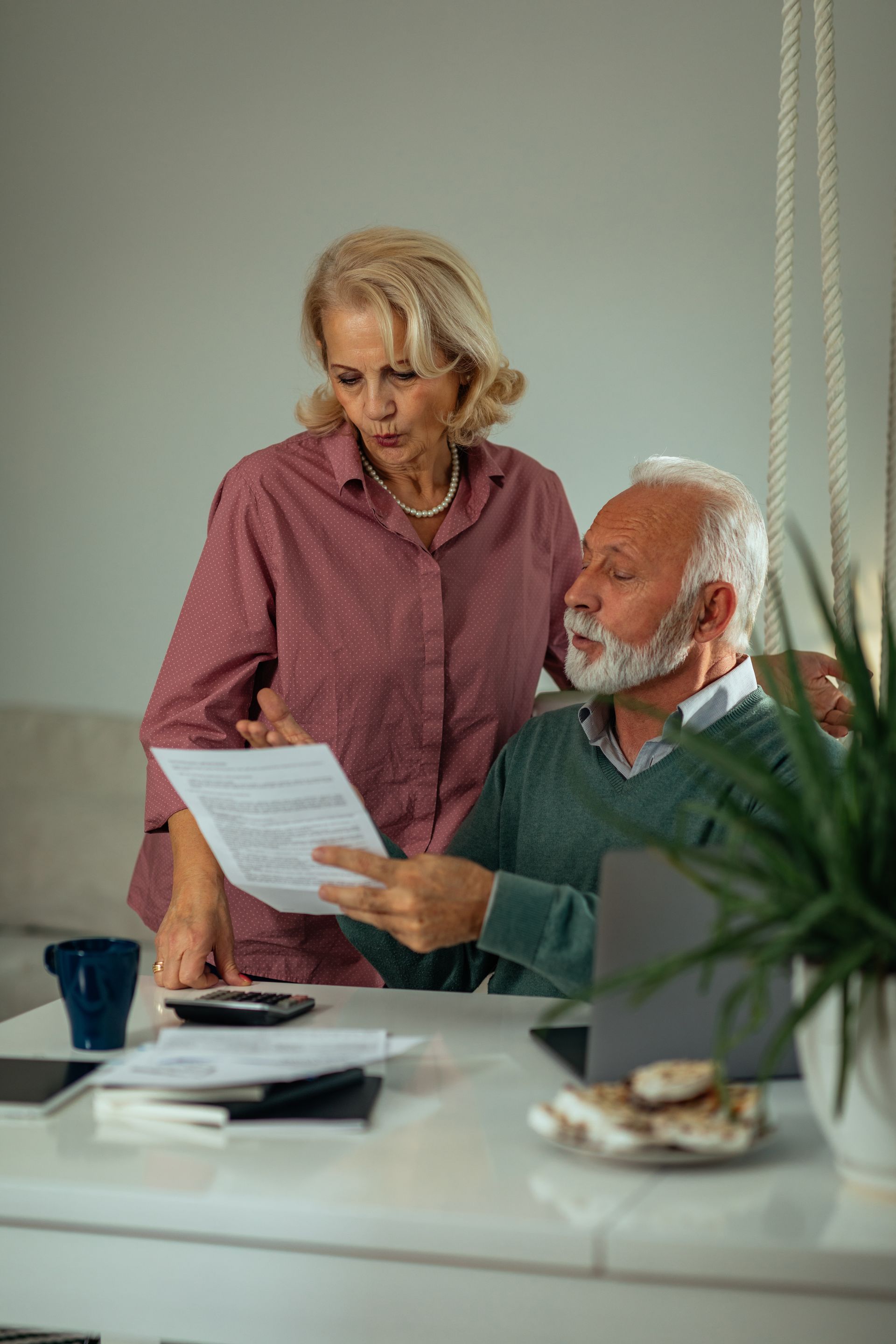 Elderly couple reviewing documents at a desk. Woman looks concerned, pointing. Man reads papers.