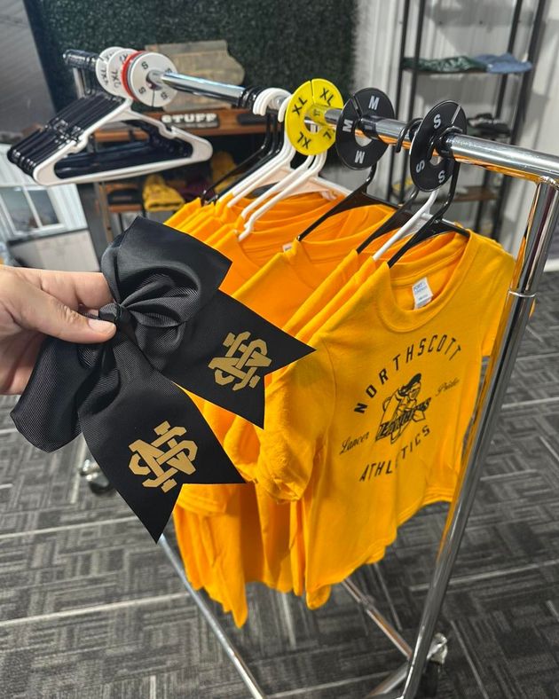 A person is holding a black bow in front of a rack of yellow shirts