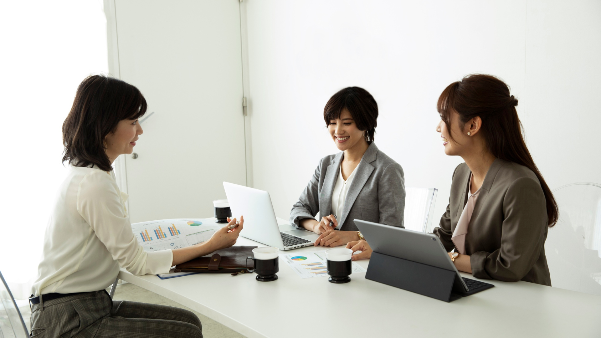 Three women in a bright office at a table; one speaking, others listen, laptop and tablet on table.
