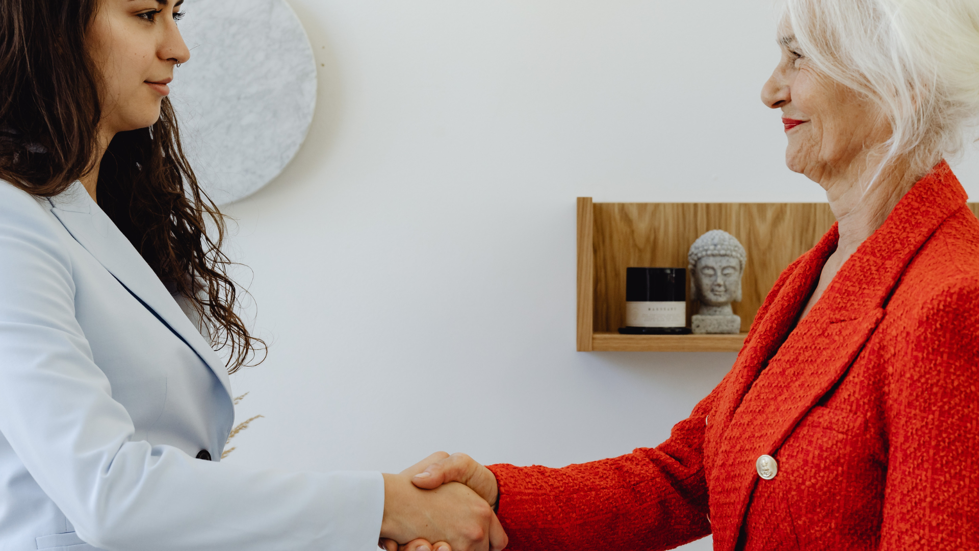 Two women shaking hands; one in a light blue blazer, the other in a red blazer. A neutral setting.