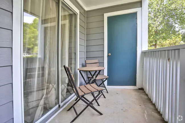 Small outdoor balcony with a table, two chairs, and a blue door. Sliding glass door on the left. Gray siding.