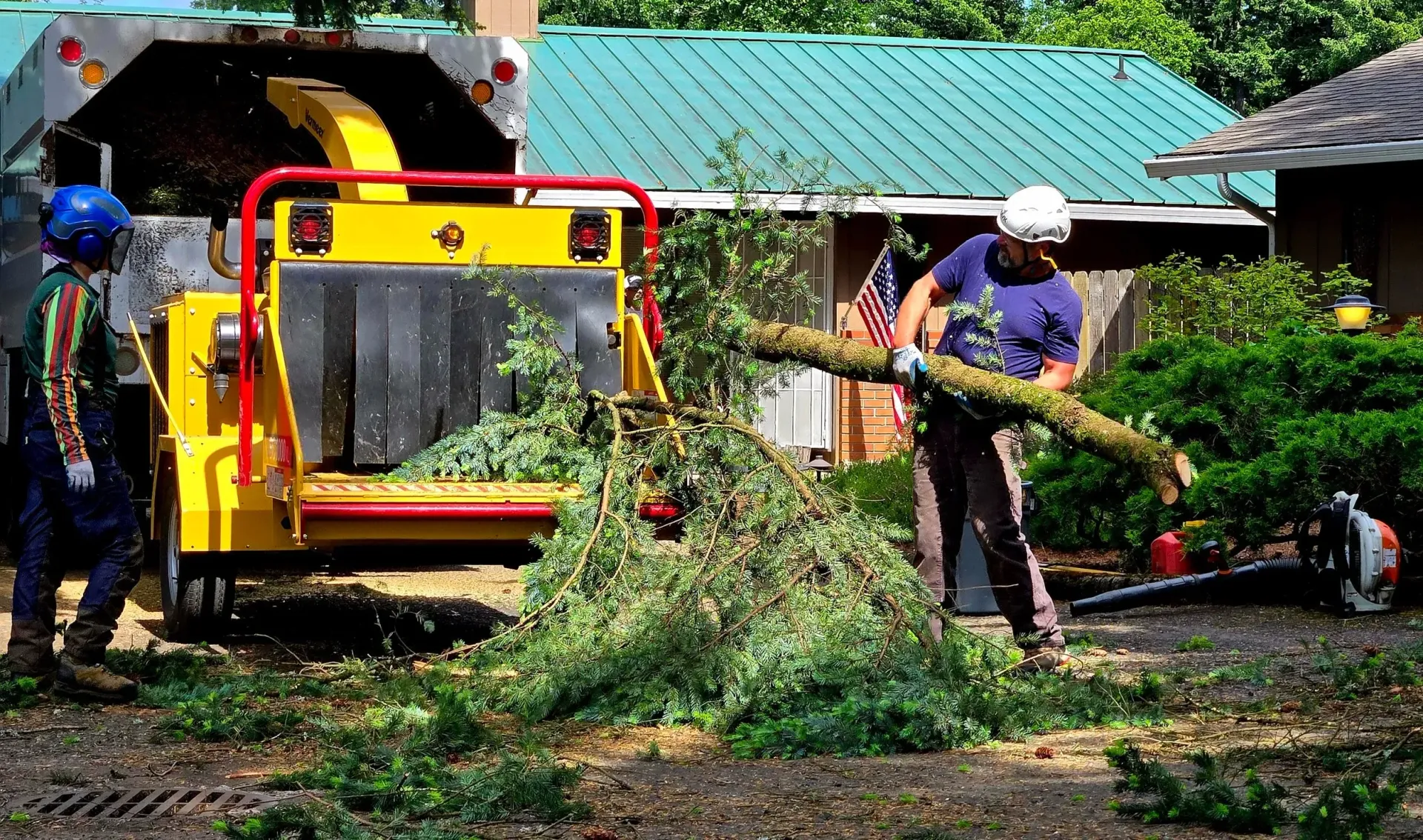 Two men are cutting a tree with a machine in front of a house.