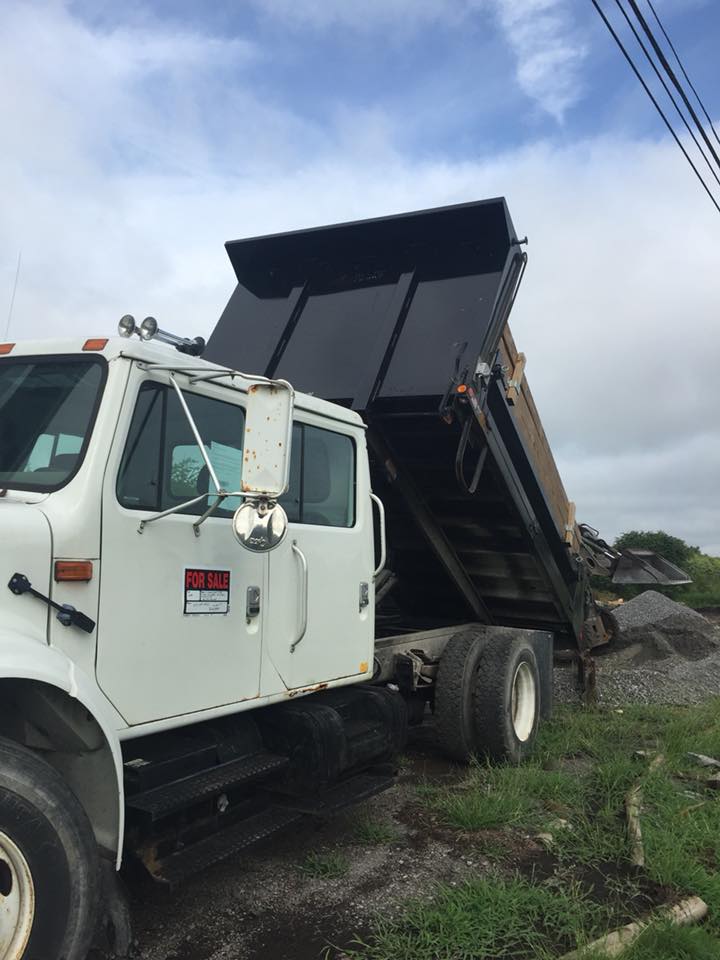 White dump truck with black bed raised, unloading gravel. Overcast sky.