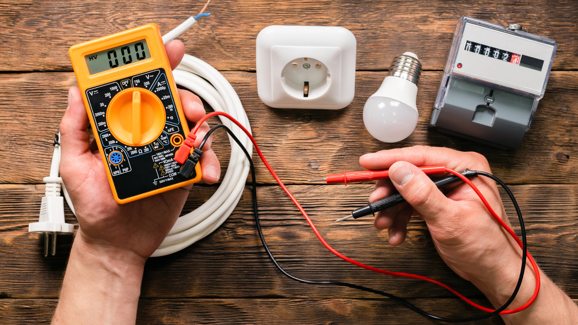 A person is using a multimeter to test a light bulb on a wooden table.