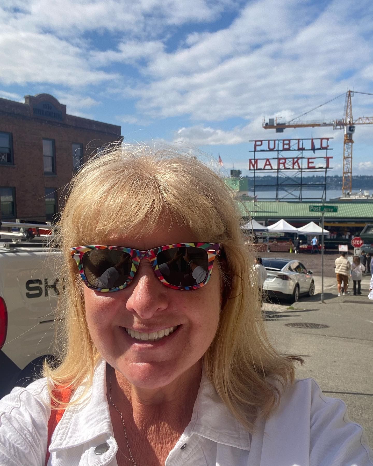 A woman wearing sunglasses is smiling in front of a sign that says public market