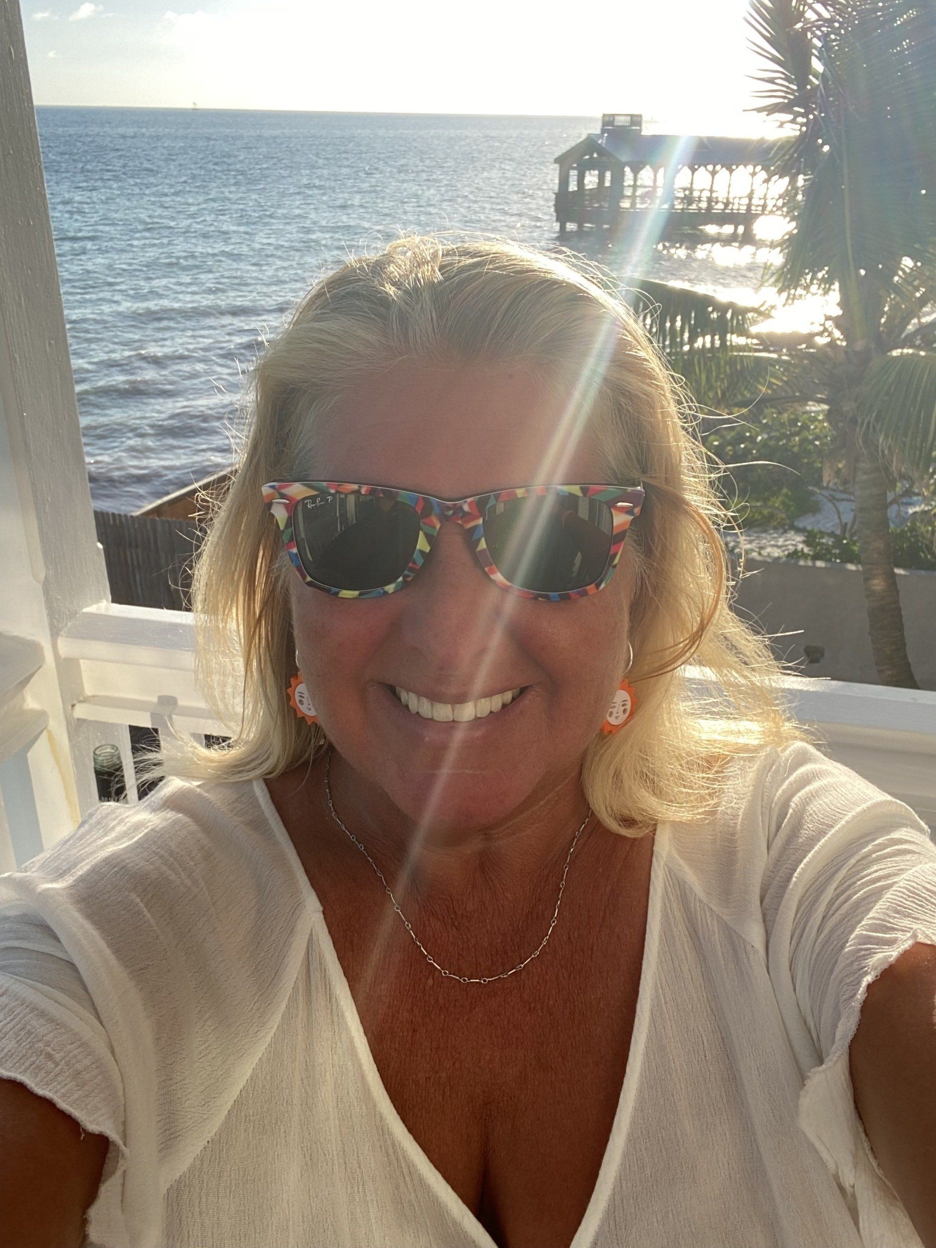 A woman wearing sunglasses is taking a selfie on a balcony overlooking the ocean.