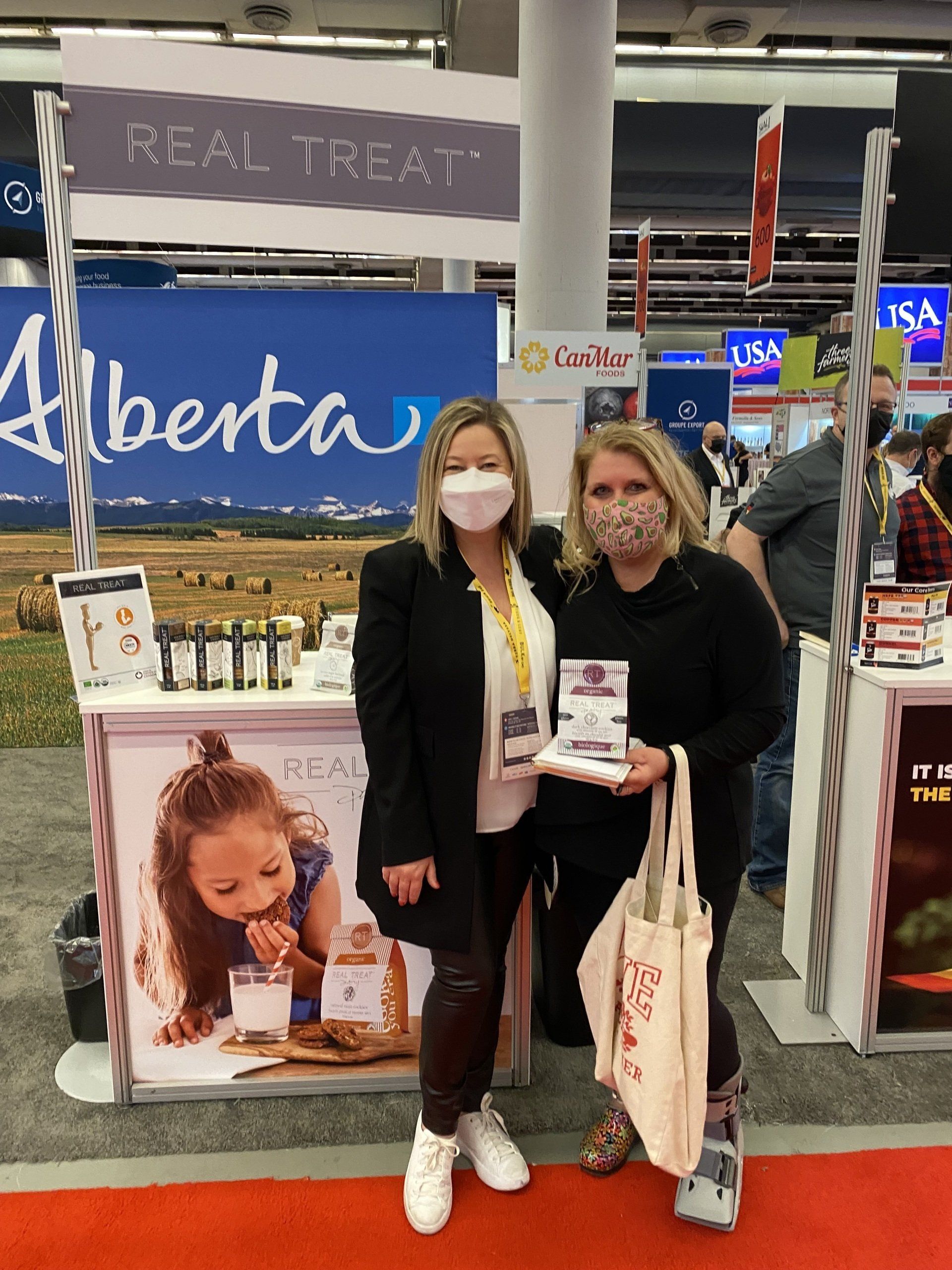 Two women wearing face masks are standing in front of a real treat alberta booth.