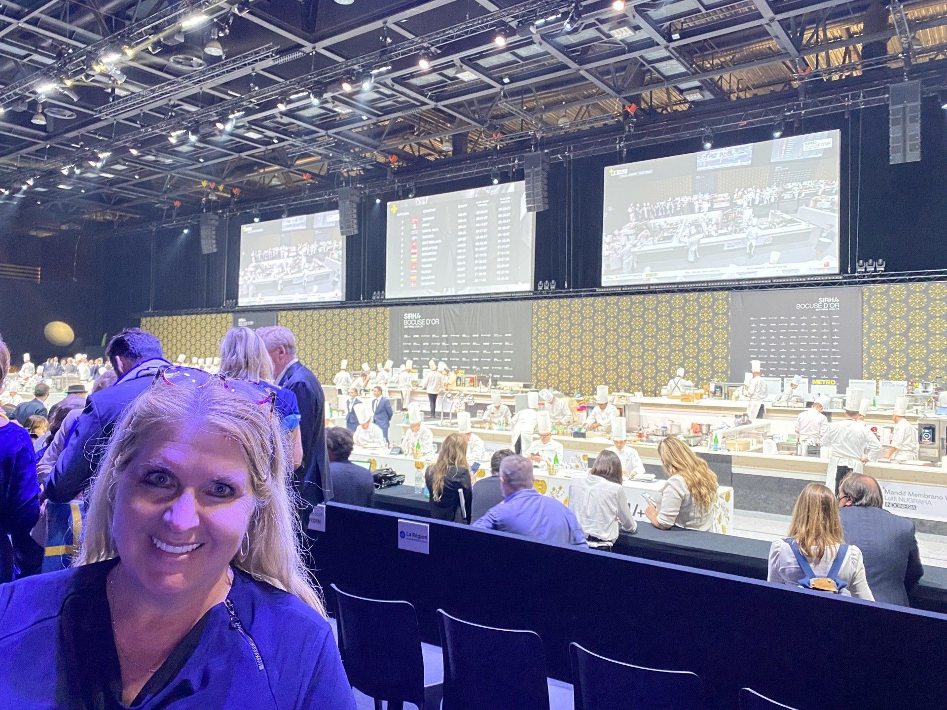 A woman is sitting in front of a large screen in a large auditorium.