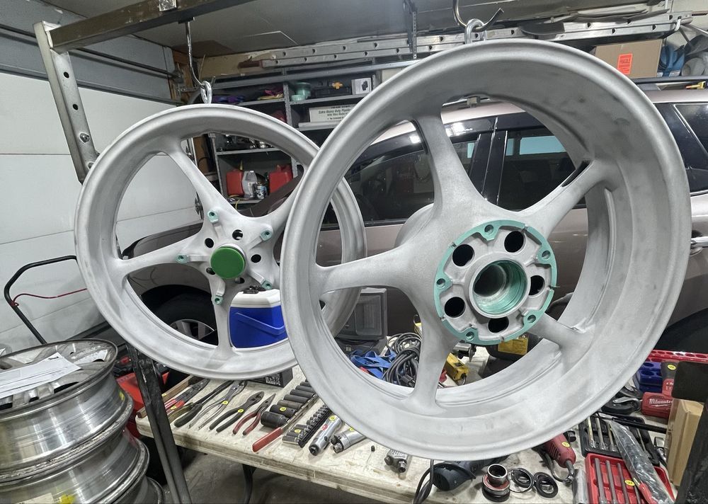 Two motorcycle wheels, light gray, hang in a garage. Tools and car are in the background.