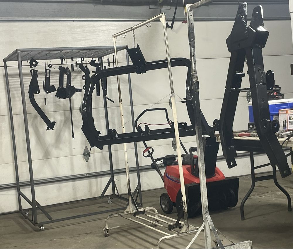 Metal automotive parts, painted black, hanging on racks in a workshop. A snowblower is visible.