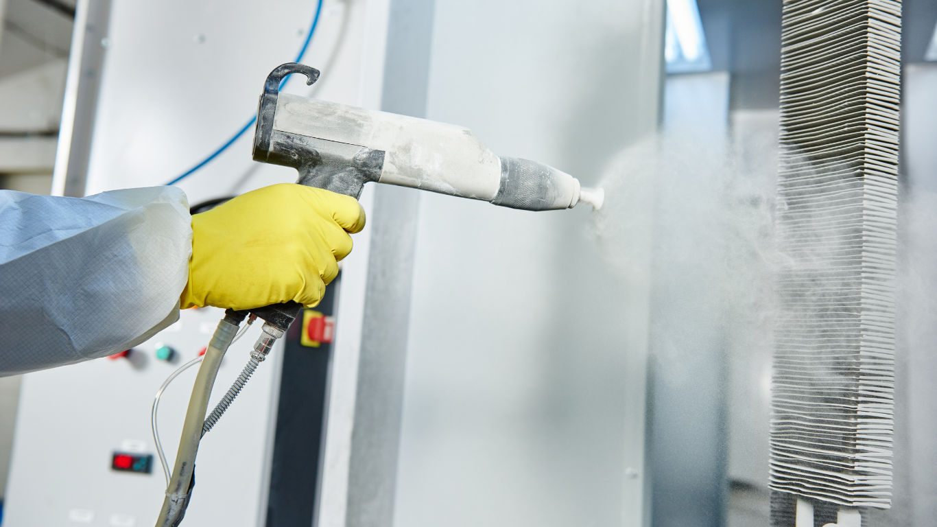 Person in yellow glove sprays powder coating on metal parts.