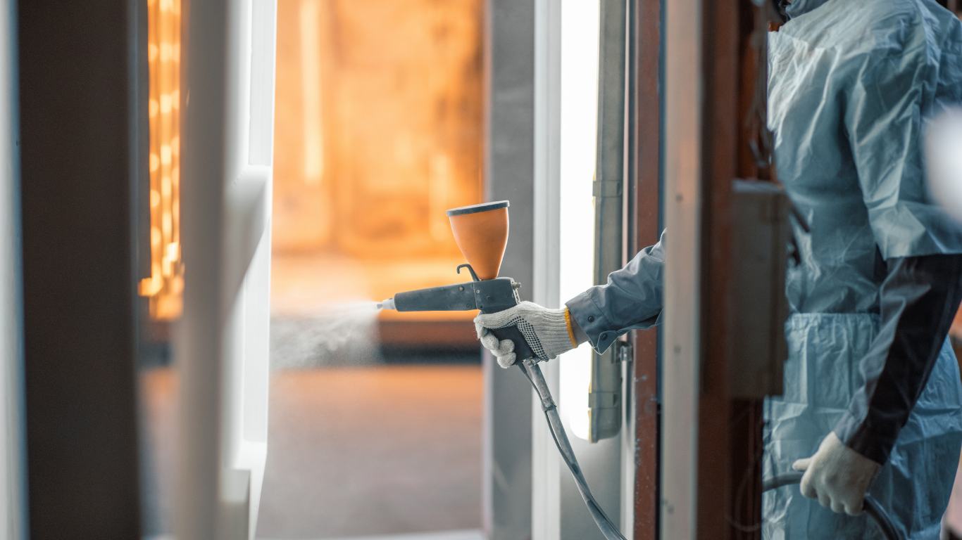 Person in protective suit spraying paint. Factory interior with white beams and orange background.