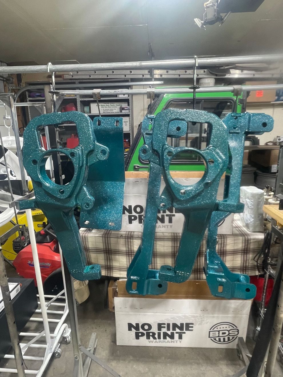 Two teal, metal automotive steering knuckles hang from a rack in a garage above a cardboard box.