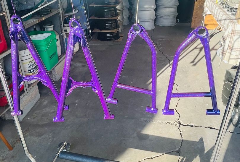 Four purple metal A-arm suspension components hang vertically on a rack in a garage or workshop setting.