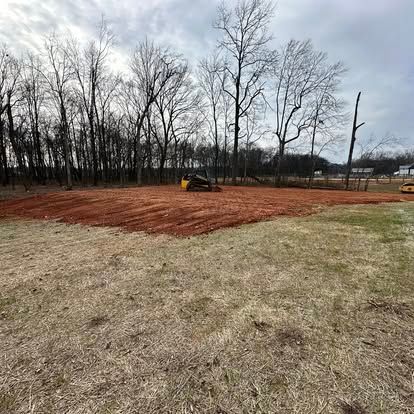 Small yellow skid steer grading a red dirt lot beside a grassy field and leafless trees under a cloudy sky