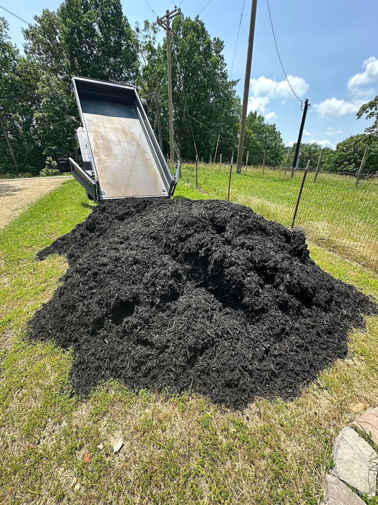 Pile of black mulch beside a tipped dump truck bed in a sunny grassy field
