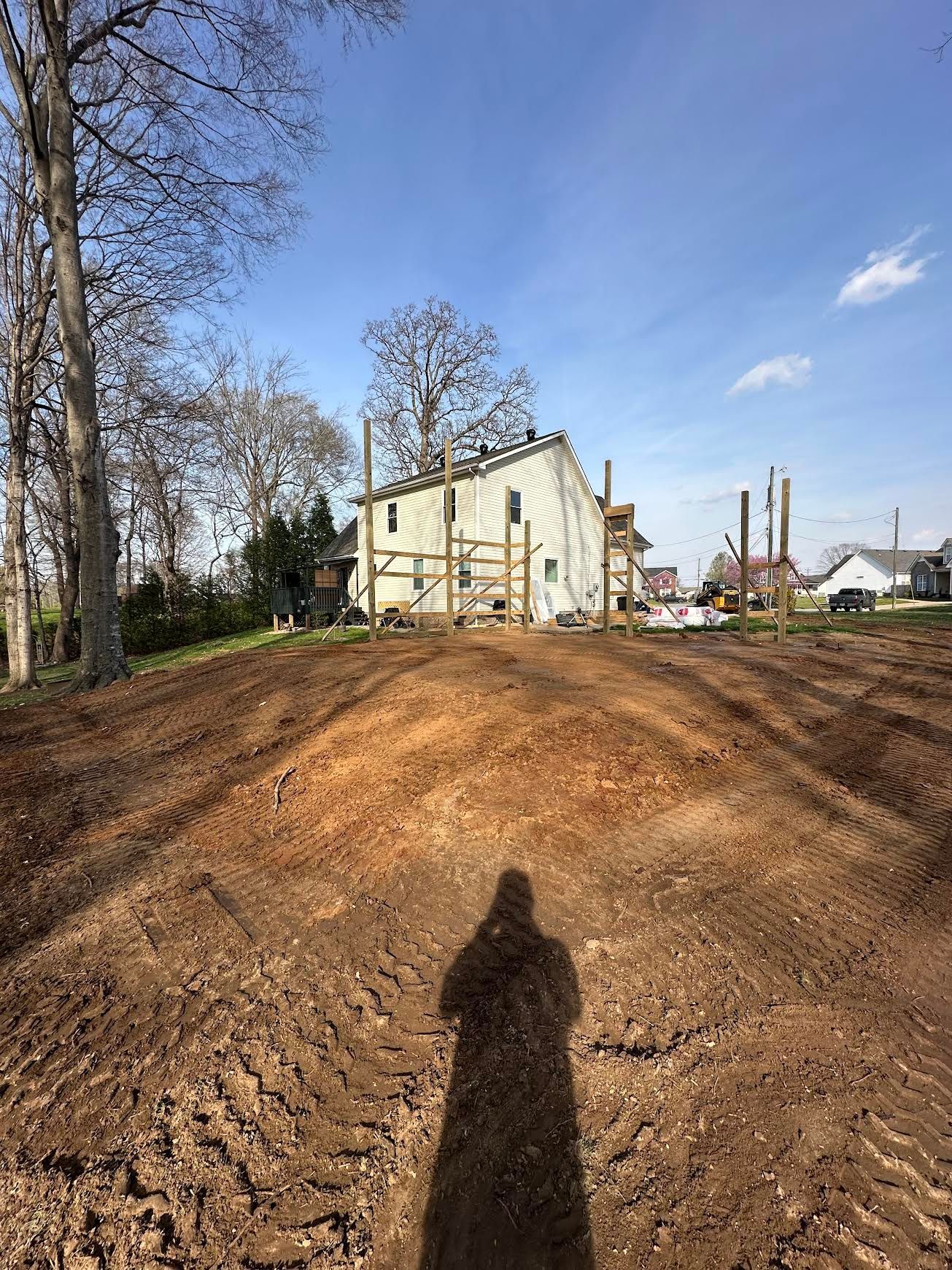 Freshly graded dirt lot with a beige house, trees, and a blue sky; a person’s shadow is in the foreground.