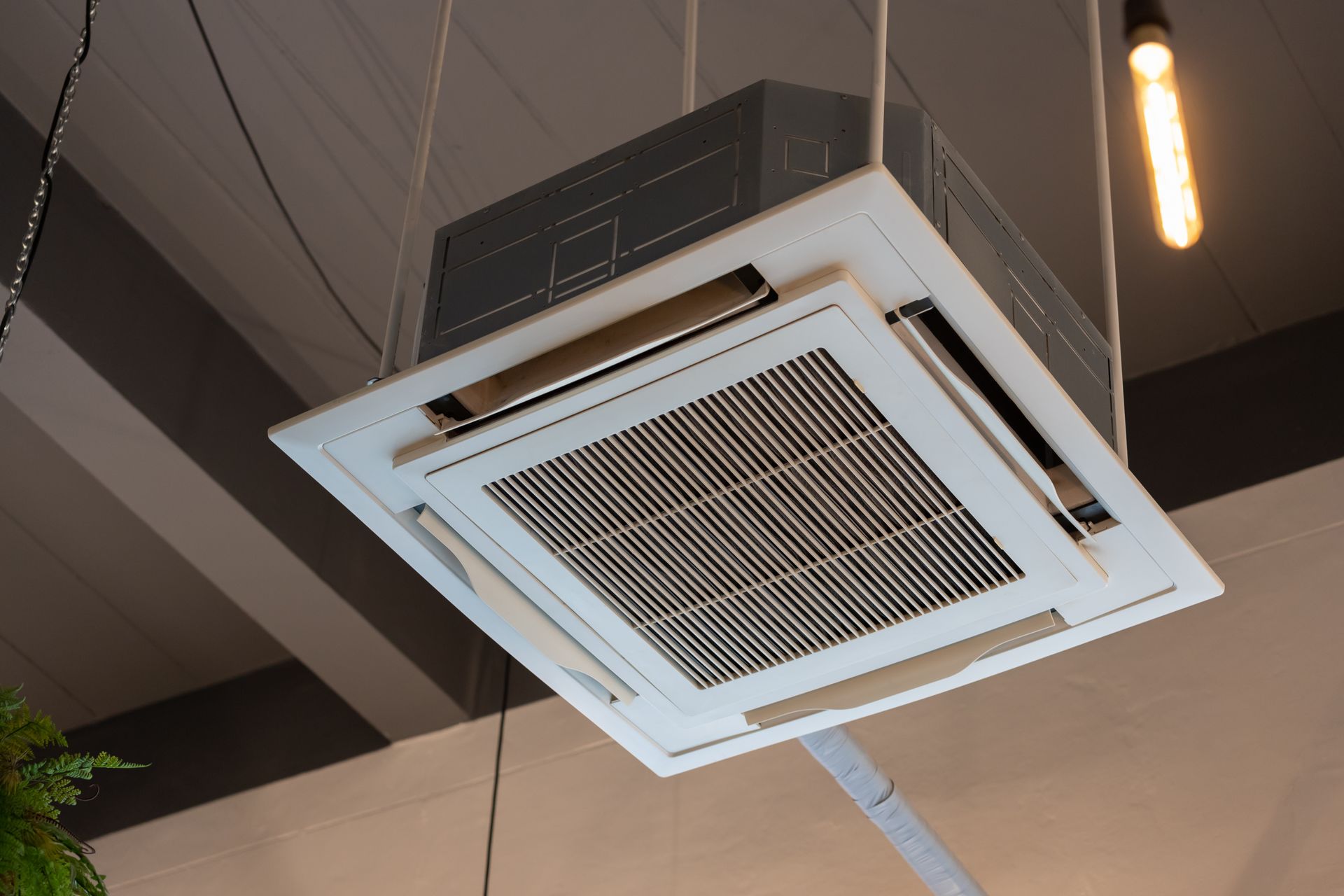 White ceiling-mounted air conditioning unit with vent slats, suspended from the ceiling.
