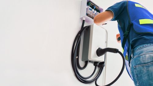 Electrician in blue vest installing an EV charger on a white wall.