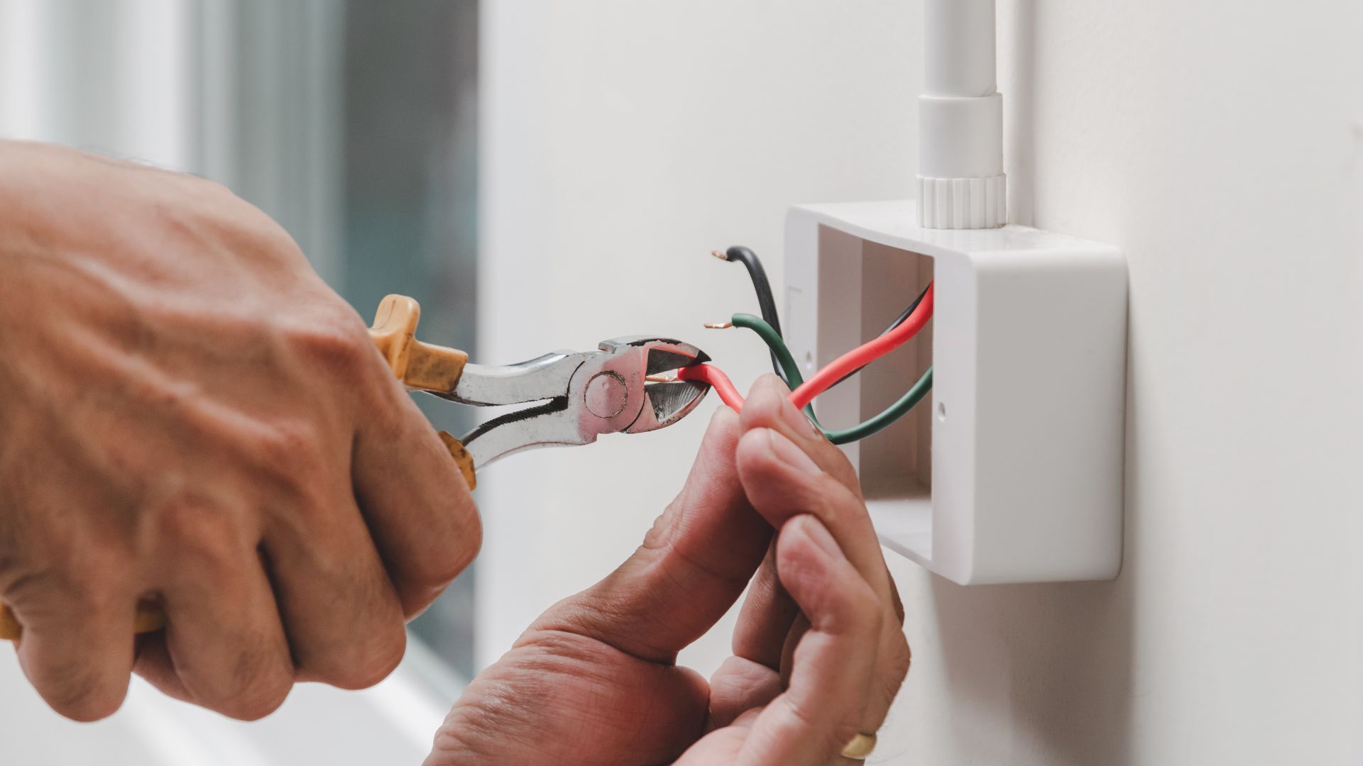 Person using pliers to cut a red electrical wire in a wall box.