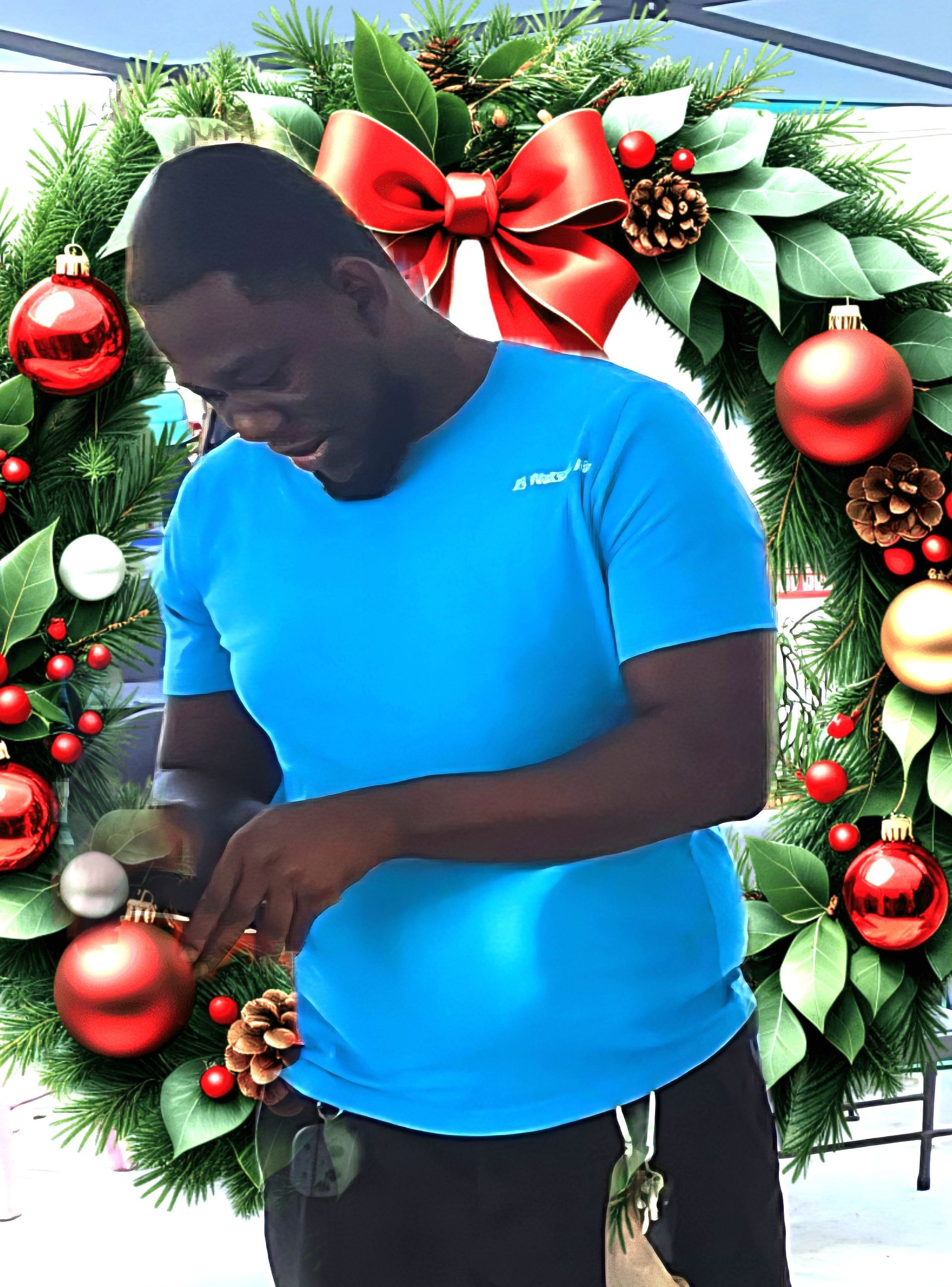 A man in a blue t-shirt standing in front of a decorative Christmas wreath adorned with red ornaments and pinecones.