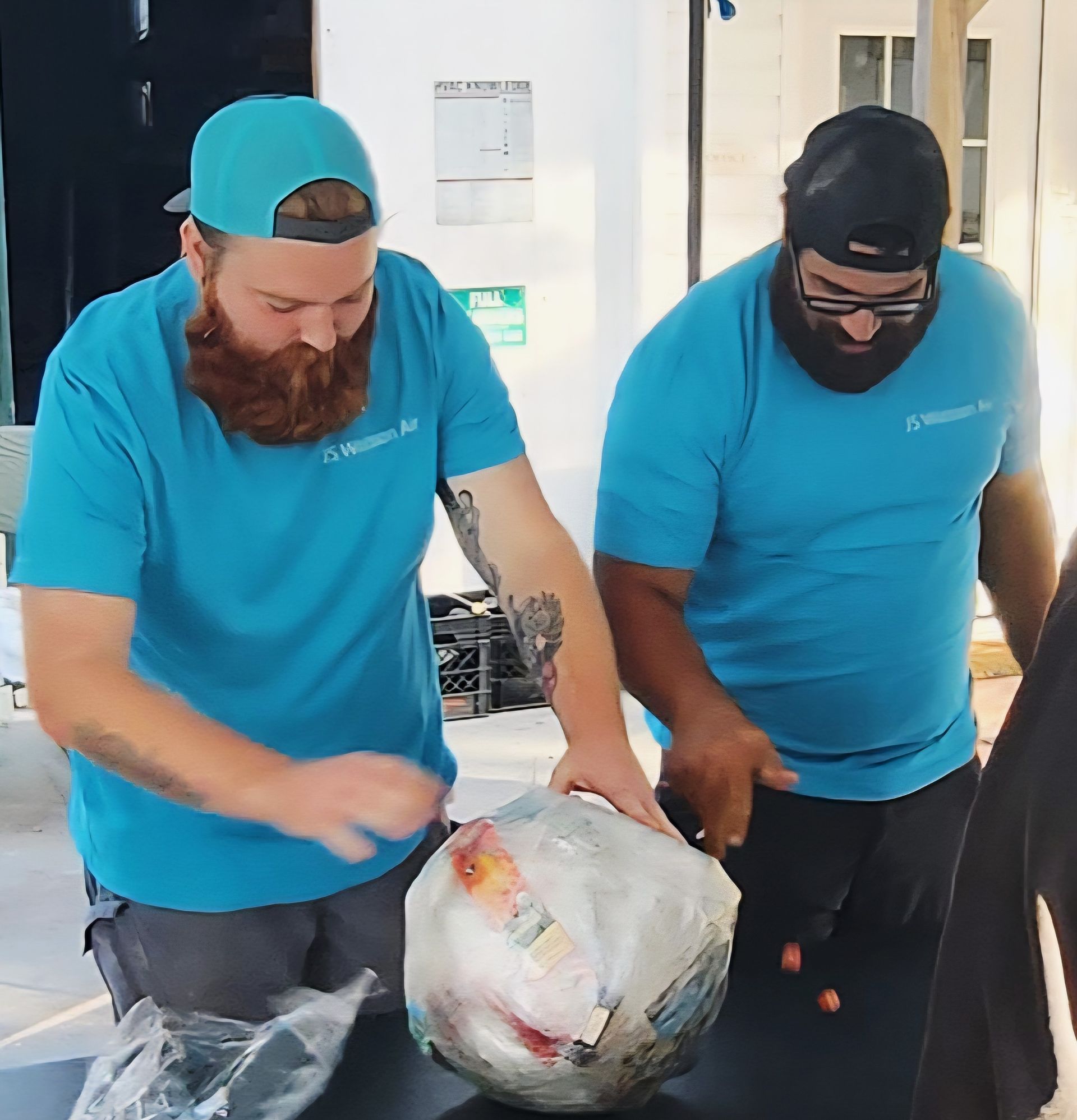 Two individuals in matching blue t-shirts wrap a round object in packing paper on a table.