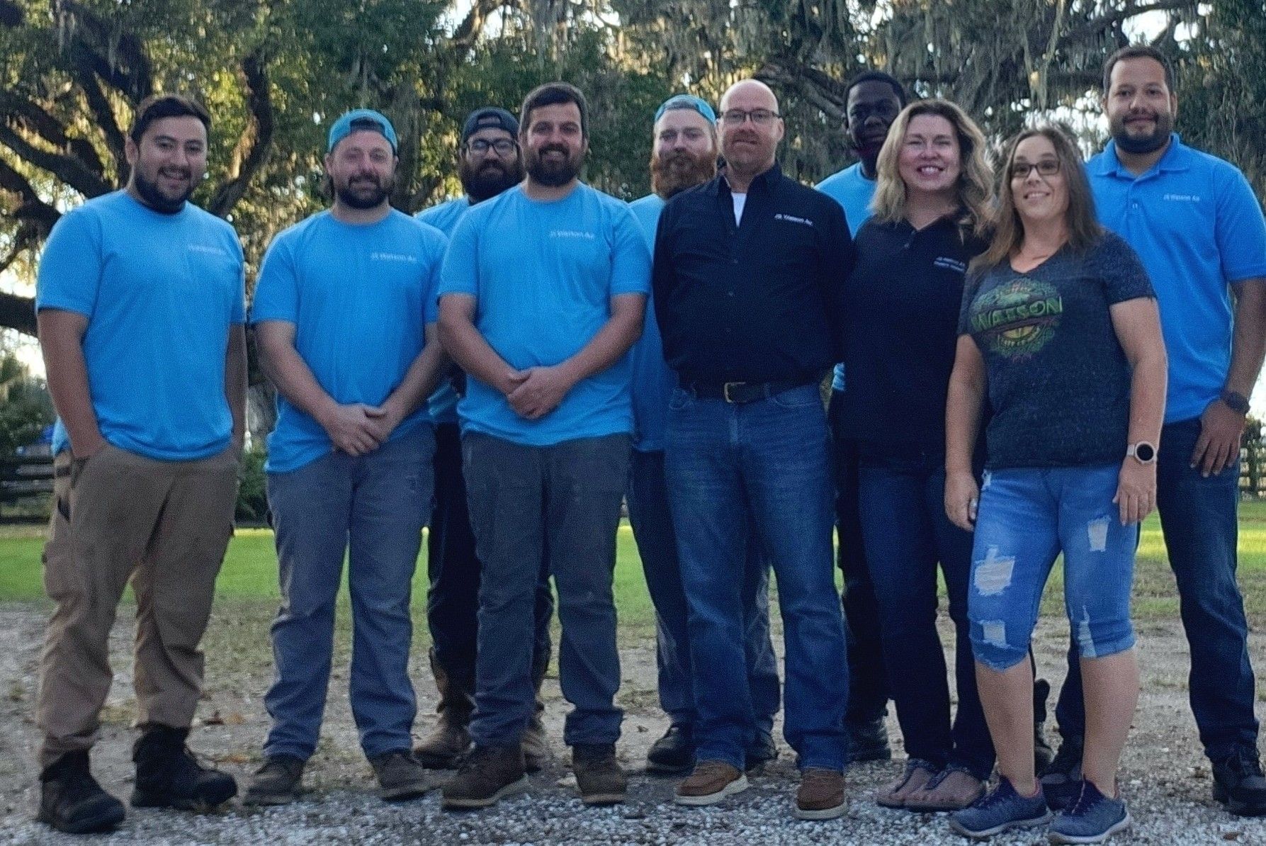 A group of nine people stand outside in a line, mostly wearing blue shirts and jeans, posing for a photo in a park setting.
