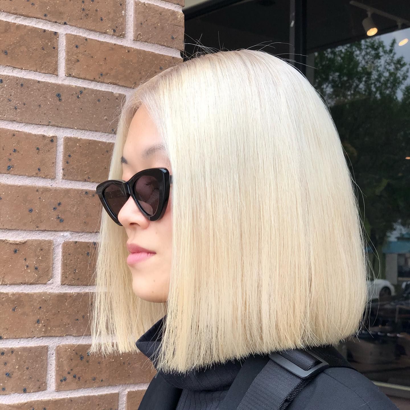 Person with blonde bob haircut, wearing sunglasses, standing near a brick wall.
