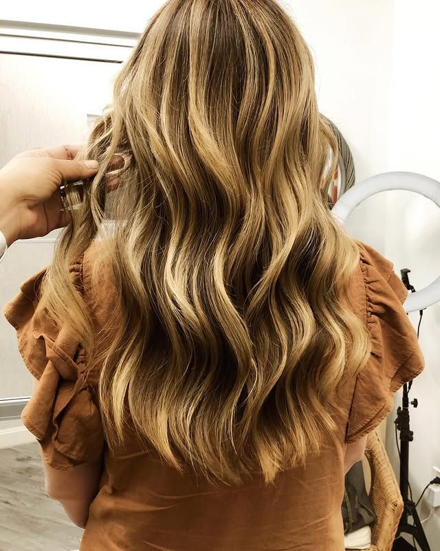 A person with long, wavy, highlighted blonde hair wearing a rust-colored ruffled top, viewed from behind.