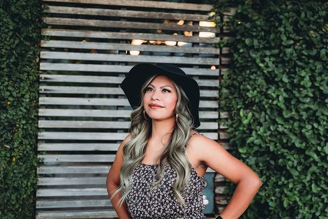 Woman in a hat and patterned dress poses in front of a wood slat wall and green vines.