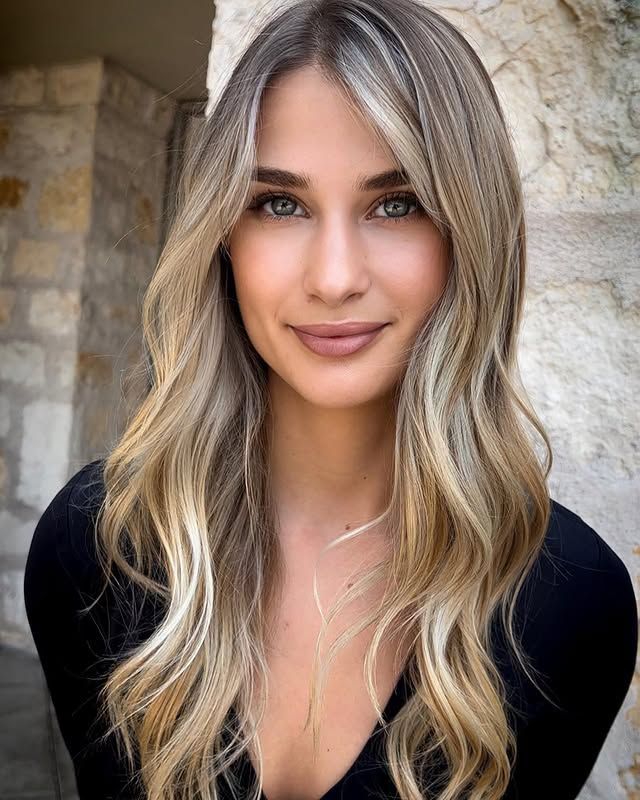 Woman with blonde-highlighted, wavy hair and a soft smile, in front of a stone wall.