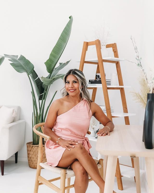 Woman in a pink one-shoulder dress sits at a wooden table in a well-lit room with plants and a wooden shelf.
