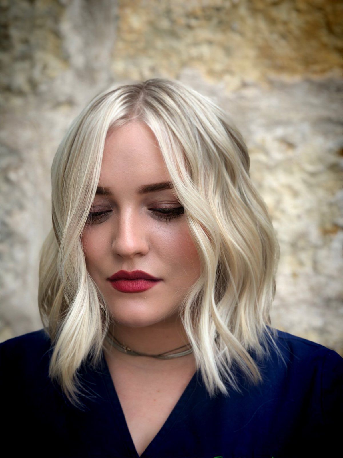Blonde-haired person with wavy bob, red lipstick, and closed eyes against a stone wall background.