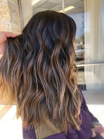 Woman's back with long, wavy, highlighted blonde hair. Person's hand adjusts a lock. Wears a rust-colored top.