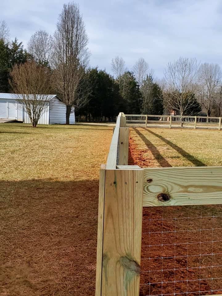 Wooden fenced paddock with wire mesh, red dirt path, winter grass, and trees under a clear sky.