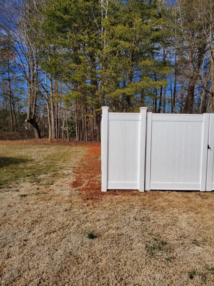 White vinyl fence panels in a grassy yard with tall trees and blue sky in the background