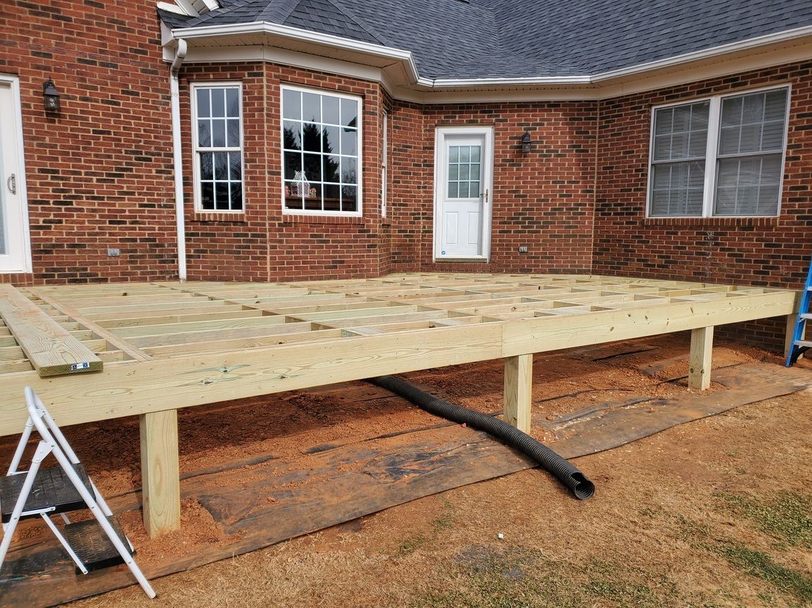 Raised wooden deck under construction in backyard beside a brick house, with ladder and support beam.