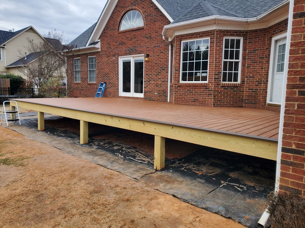 Raised wooden deck attached to a brick house, with concrete support posts and unfinished yard below