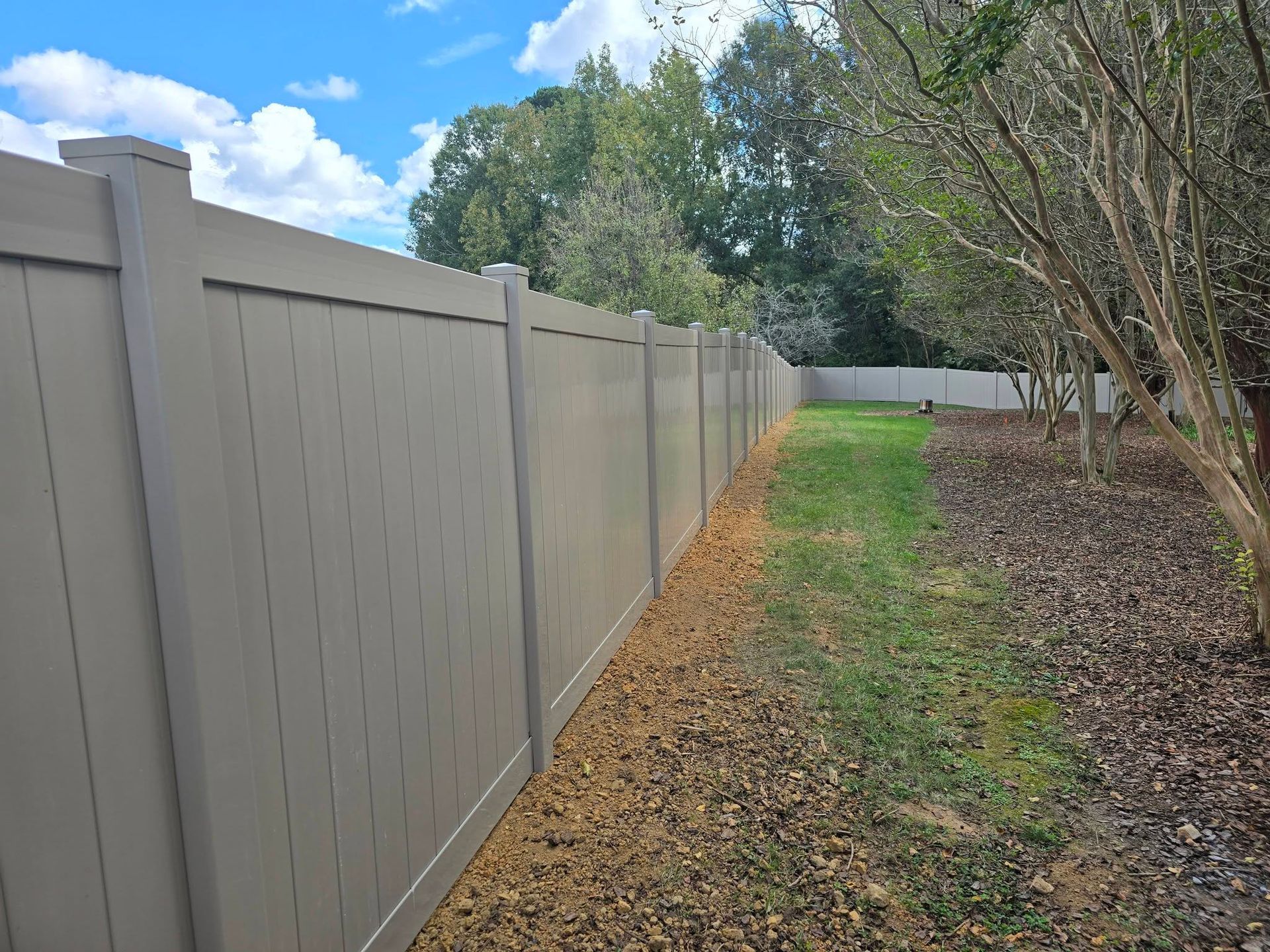 Gray vinyl fence along a narrow grassy yard with trees and a partly cloudy sky