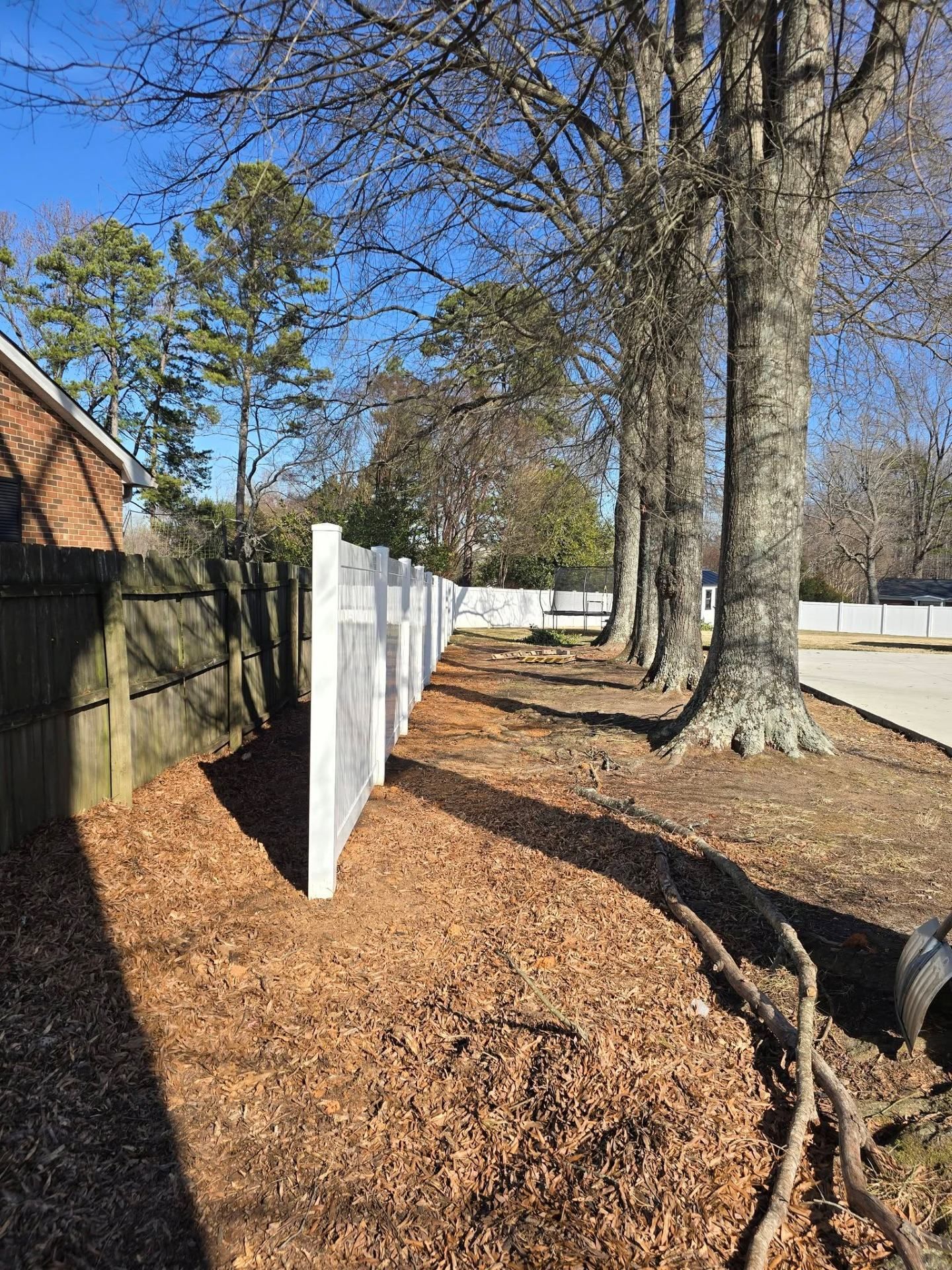 White fence posts along a mulched yard beside tall trees on a sunny day
