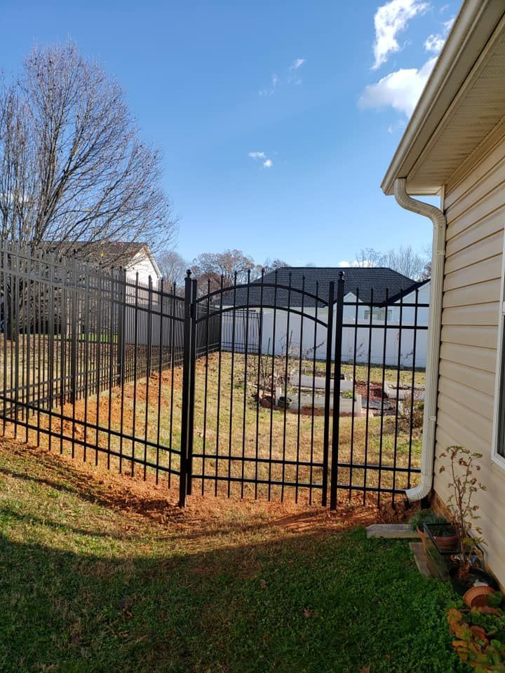 Black metal gate beside a beige house, opening to a small fenced yard under a bright blue sky.