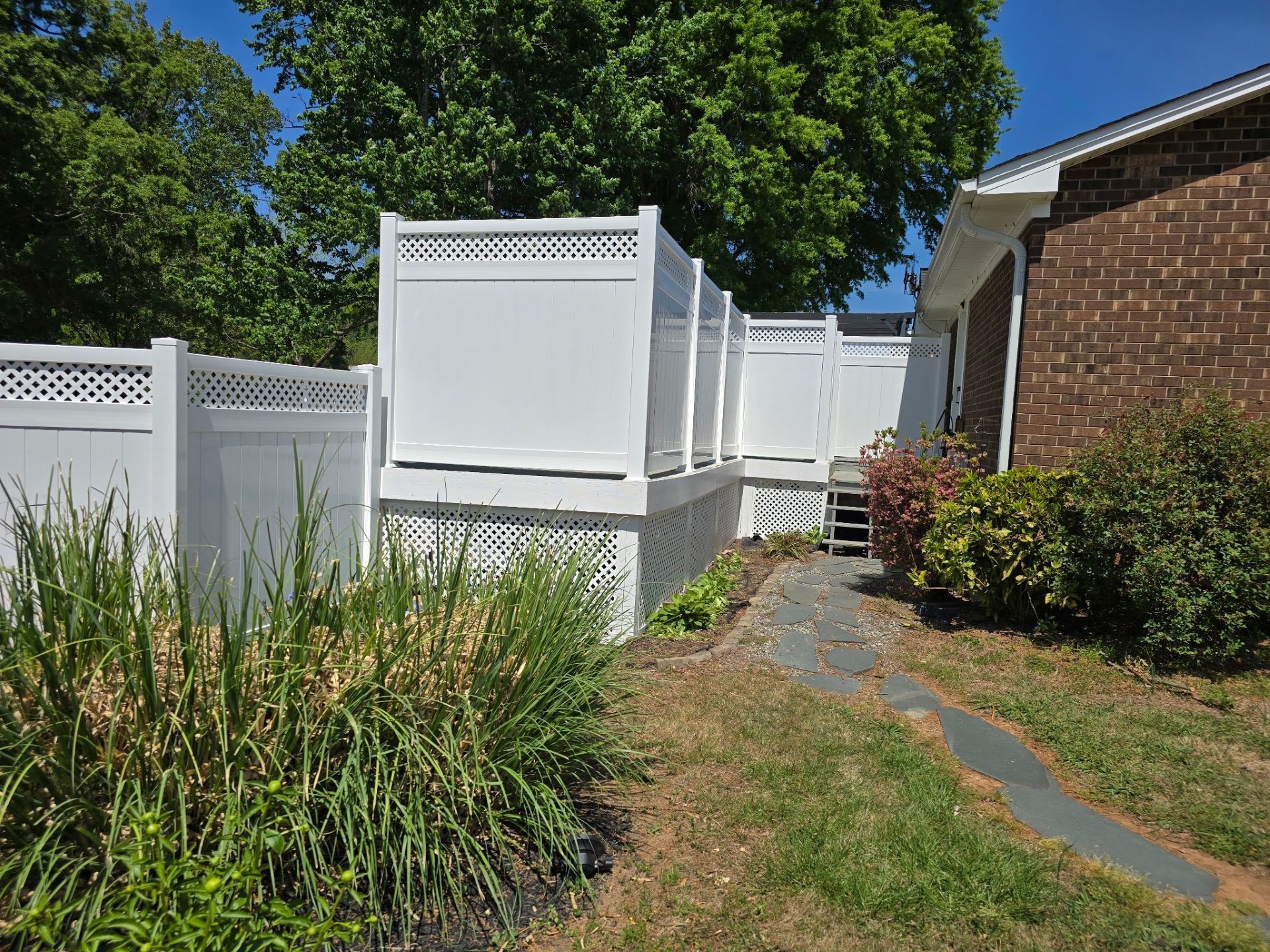 Backyard path beside white fence, brick house, and tall green plants under a sunny tree.